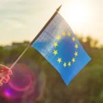 Hand holds European Union flag in an open window. Background blue sky, silhouette of the city